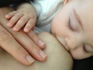 baby with eyes closed, holding mother's hand while feeding at breast